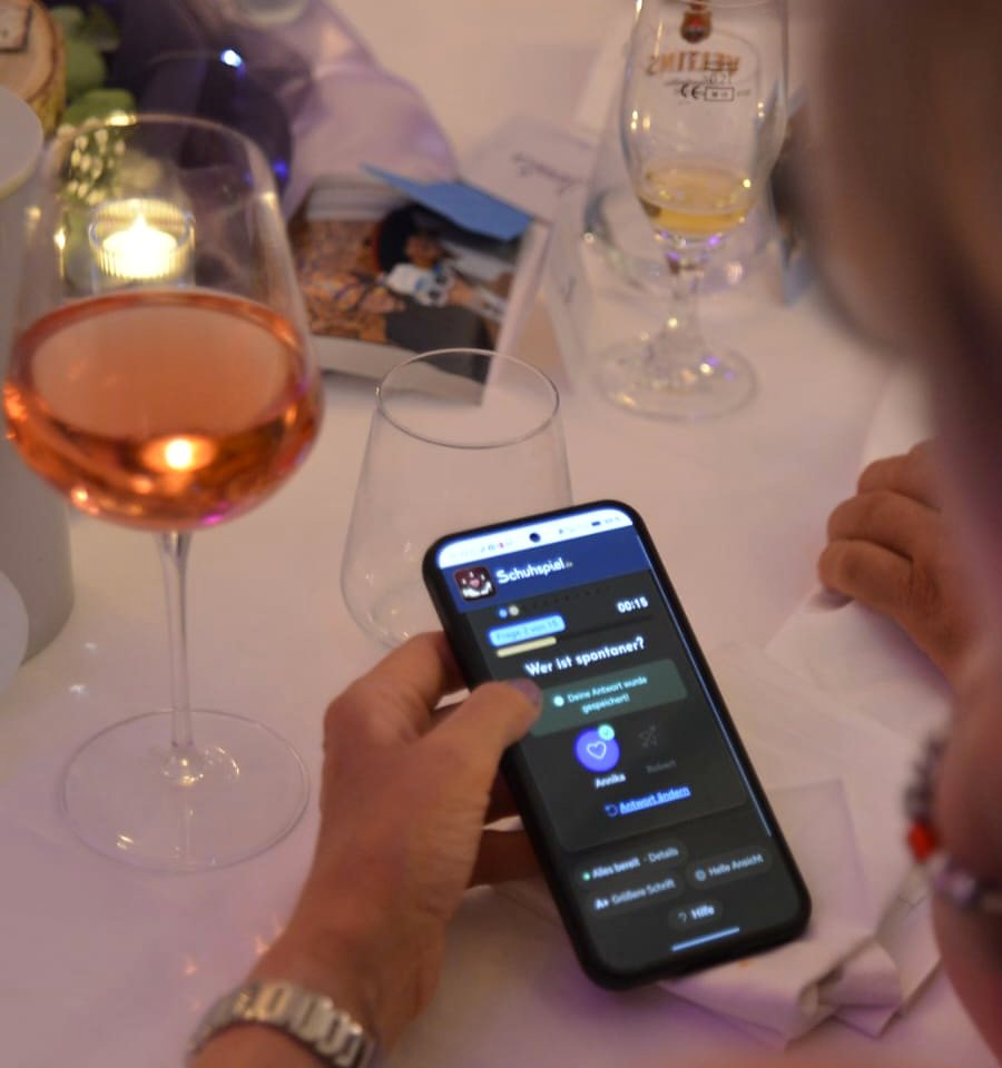 Gast beantwortet die Schuhspiel-Frage am eigenen Smartphone, daneben ein Glas Wein auf der festlich gedeckten Tafel.