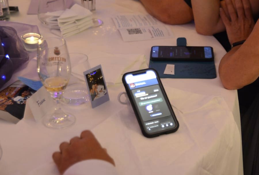 Zwei Smartphones mit der Schuhspiel-Ansicht und die ausgedruckte Beitrittskarte liegen auf einem Hochzeitstisch.